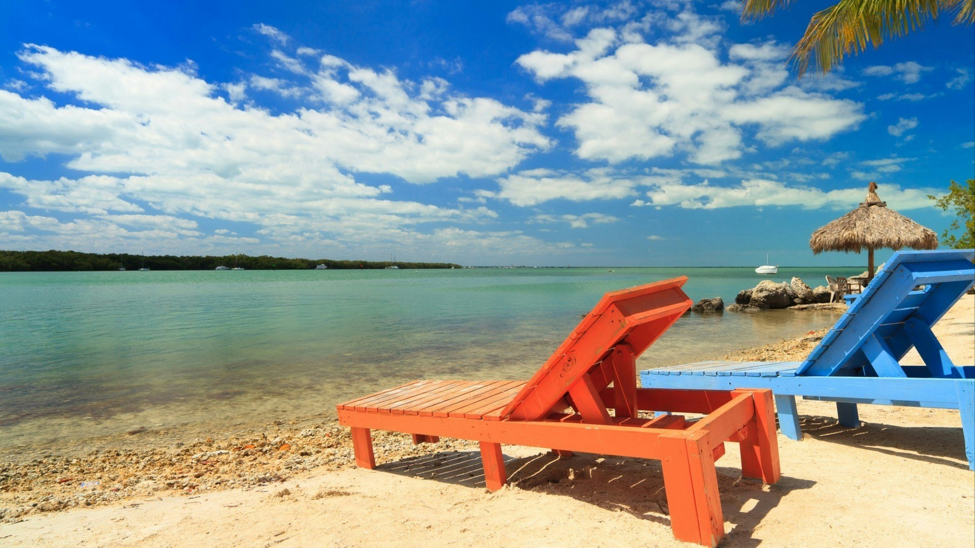 Wooden beach lounge chairs along the shoreline of the Florida Keys with pretty blue sky and clouds and thatched hut in the background.